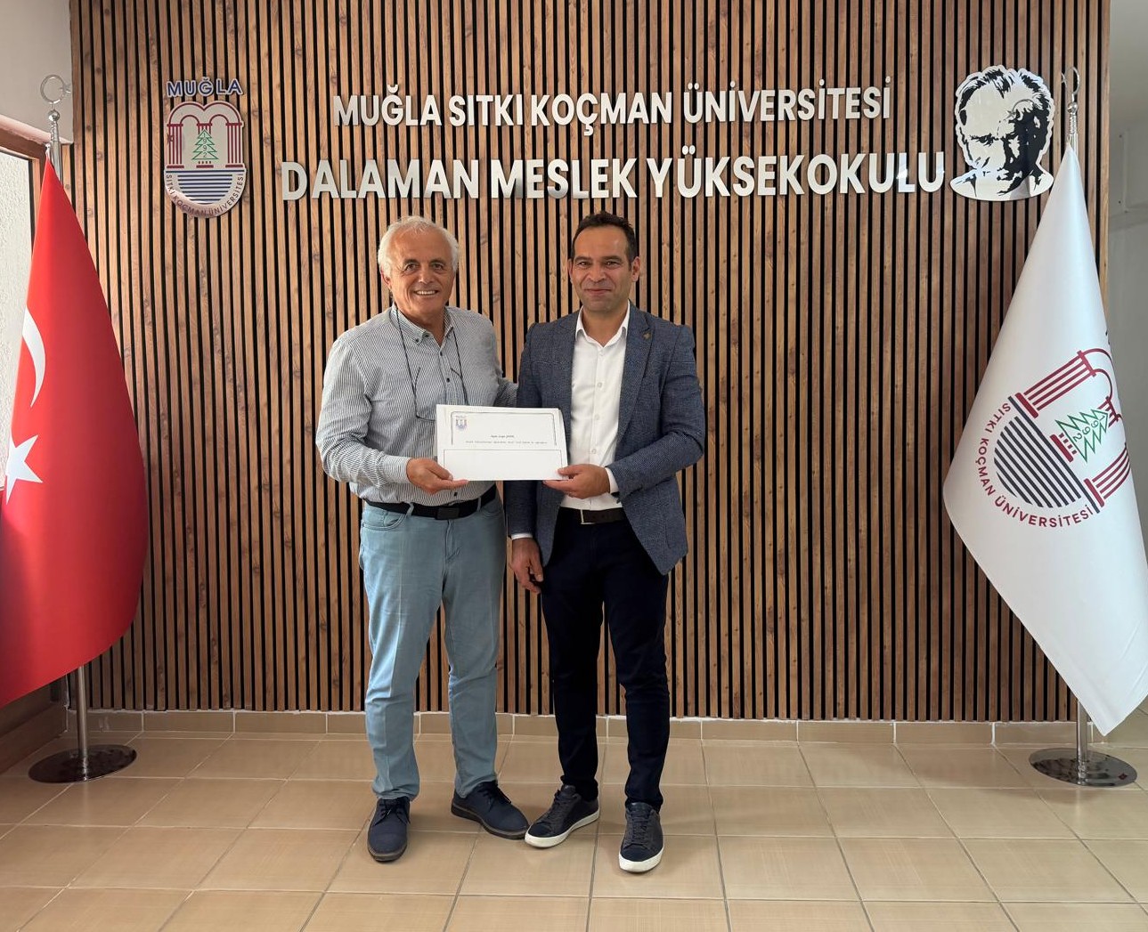 Cengiz Şatır ve Doç. Dr. Ozan Kaya, Dalaman Meslek Yüksekokulu'nun logolu duvarının önünde birlikte bir belge tutarken görülüyor. Bu kare kurumlar arası iş birliğini simgeleyen bir kapanış pozu niteliğinde.